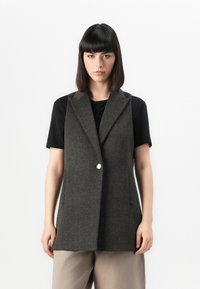 Vest gri din lână cu rever ascuțite, închidere cu un singur buton și buzunare laterale, purtat peste un tricou negru, asociat cu pantaloni bej.