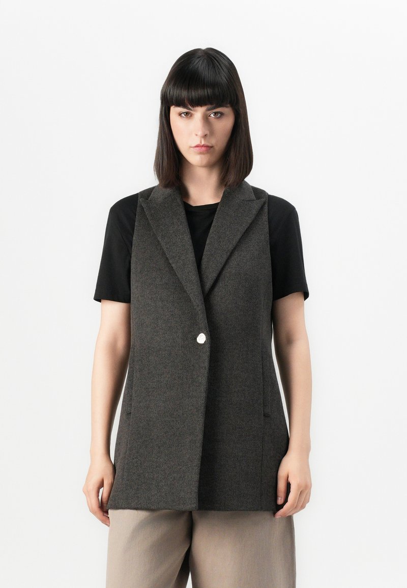 Vest gri din lână cu rever ascuțite, închidere cu un singur buton și buzunare laterale, purtat peste un tricou negru, asociat cu pantaloni bej.