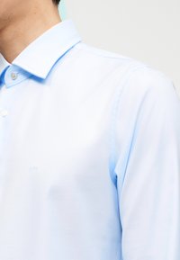 Άνδρας που φοράει ανοιχτό μπλε πουκάμισο με κουμπιά και κλασικό γιακά, εμφανίζεται από τον λαιμό έως τον άνω βραχίονα πάνω σε απλό φόντο.