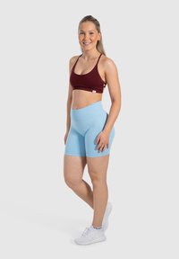 Reggiseno sportivo color mogano e pantaloni corti a vita alta blu chiaro. Modella con scarpe da ginnastica bianche. Tessuto liscio ed elastico con branding minimo.
