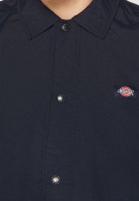 Close-up of a black collared shirt with black snap buttons and a small rectangular Dickies logo patch on the chest.