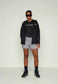 Frau in schwarzer Terrex-Jacke und T-Shirt, grauen Shorts, schwarzen Socken, schwarzen Turnschuhen und großen schwarzen Sonnenbrillen, vor einem schlichten Hintergrund stehend.
