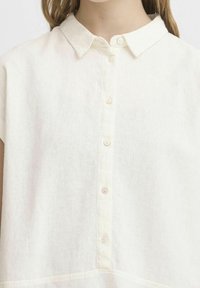 Primo piano di una persona che indossa una camicia di lino bianca con colletto a punta e una fila verticale di bottoni sul davanti.
