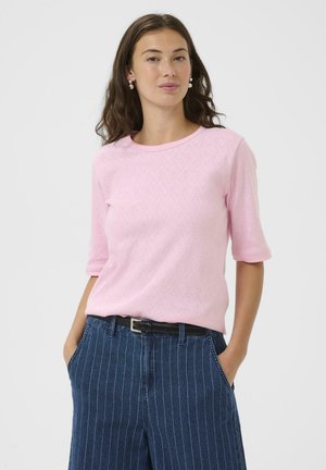 Femme aux cheveux bruns portant un haut texturé à manches courtes rose clair et un jean bleu à fines rayures, les mains dans les poches.