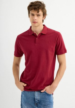 Camisa polo roja de tejido suave, con cuello, mangas cortas y un pequeño logo en el lado izquierdo del pecho. Combinada con pantalones cortos de mezclilla azules.