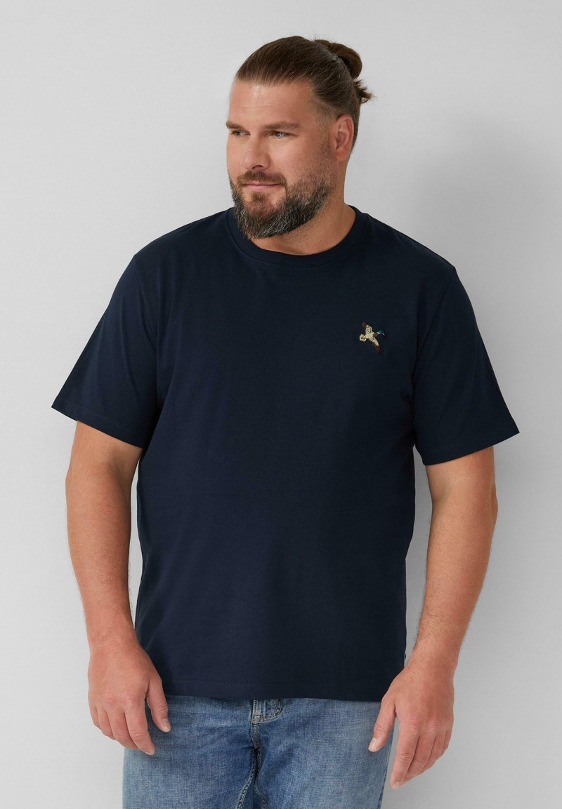 Man met baard en samengebonden haar, gekleed in een marineblauwe t-shirt met een klein vogel logo en lichtblauwe jeans, staande tegen een effen muur.