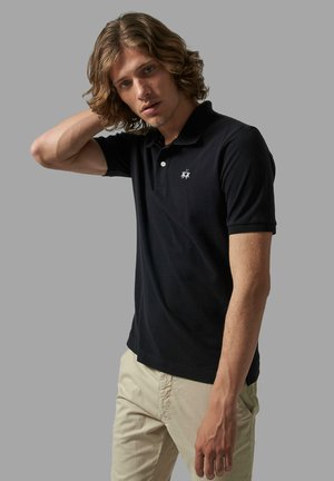 Polo shirt nera con apertura a due bottoni e logo ricamato sul petto. Tessuto morbido con maniche corte, abbinato a pantaloni color khaki chiaro.