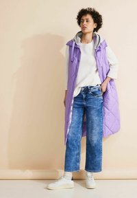 Lila quiltad pufferväst med grå huva, tillsammans med ljusblå vida jeans och vita sneakers. Slät textur och subtil glans.