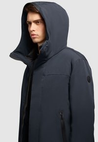 Mørkeblå pufferjakke med stor hætte, lynlås foran og sidelommer. Har en glat, mat tekstur og et logo-patch på ærmet.