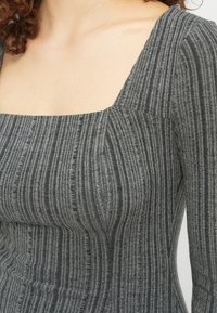 Γυναίκα που φοράει εφαρμοστή γκρι πλεκτή μπλούζα με μακριά μανίκια και τετράγωνη λαιμόκοψη, εμφανίζοντας τον πάνω κορμό και τον λαιμό.