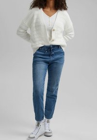 Vit stickad kofta med öppen front, utrustad med två knappar, parat med blå jeans och vita högkantiga sneakers. Texturerat stickat mönster synligt.