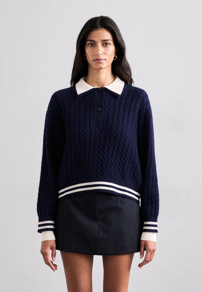 Pulover tricotat bleumarin cu un model texturat de cablu, guler alb și tiv cu dungi ribbed. Patru butoane negre în față îmbunătățesc designul.