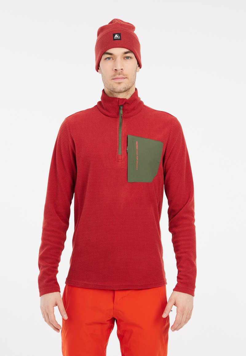 Rode getextureerde pullover met een kwartrits, groene borstzak en een bijpassende rode beanie, gedragen met feloranje broeken.