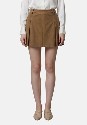 Donna che indossa una camicetta bianca a maniche lunghe, gonna marrone plissettata in pelle scamosciata e mocassini beige a punta, in piedi davanti a uno sfondo semplice.