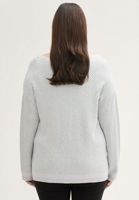 Hellgrauer Pullover mit Fischgrätmuster, weit geschnittenen Ärmeln und einem leicht verlängerten Saum. Der Stoff wirkt weich mit einer strukturierten Oberfläche.