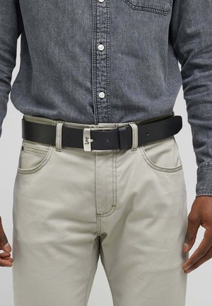 Schwarzer Leder Gürtel mit silberner Metallschnalle, getragen über hellgrauen Hosen, mit einem blauem Jeanshemd, das teilweise sichtbar ist.