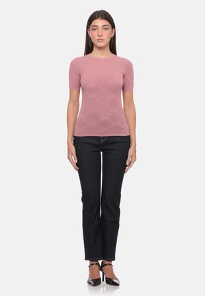 Donna con una maglietta aderente rosa a maniche corte, jeans scuri a gamba dritta e scarpe nere con tacco alto, in piedi con le braccia lungo i fianchi.