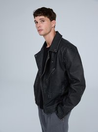 Giacca da moto in pelle nera con ampio colletto, tasche con cerniera e dettagli di cuciture a contrasto, indossata sopra una maglia scura e pantaloni grigi.