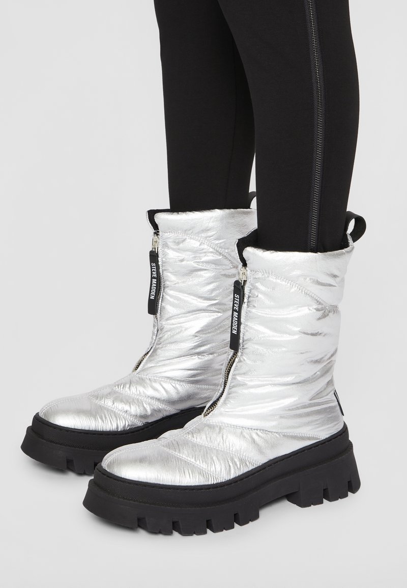 Srebrne, ocieplane buty z połyskującą, pikowaną teksturą, wyposażone w dwie czarne zamki błyskawiczne i masywne czarne gumowe podeszwy. Widoczne czarne legginsy.