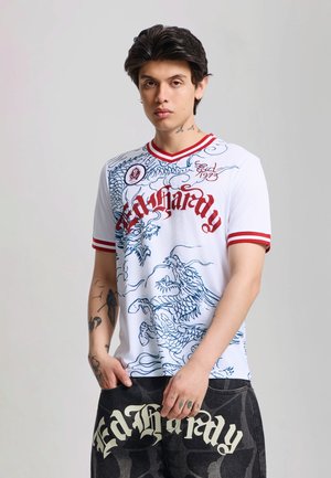 Giovane uomo con capelli scuri che indossa una camicia bianca con scritte rosse e motivo di drago blu, e pantaloni scuri con testo bianco in grassetto.