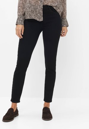Vrouw met zwarte skinny broek, een bruin gebloemde blouse en donkerbruine loafers, staand tegen een effen witte achtergrond.