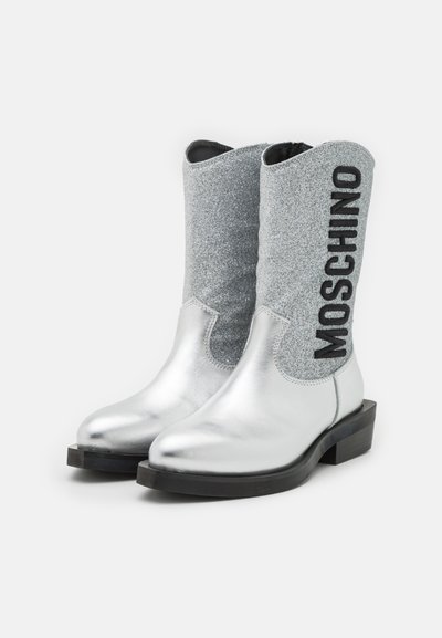 Bottes de cowboy argentées avec une finition métallique et un dessus pailleté, présentant un logo "MOSCHINO" noir et un petit talon épais.