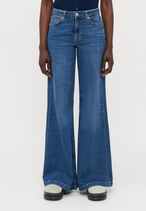 Wijde blauwe denim jeans met een hoge taille, uitgerust met vijf zakken, contrasterende stiksels en een gladde textuur. Draagt met groene schoenen.