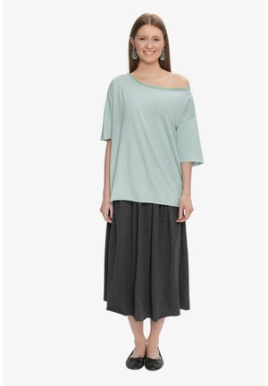 Kobieta uśmiechnięta, ubrana w oversize jasnoniebieską bluzkę odsłaniającą ramiona, ciemnoszarą spódnicę midi, czarne baleriny i duże kolczyki, stojąca na białym tle.