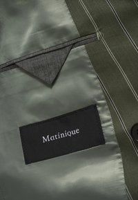 Ένα πράσινο ύφασμα με σατινέ φινίρισμα διαθέτει μια μαύρη ετικέτα με την ένδειξη "Matinique." Είναι ορατή μια γκρι τσέπη με αντίθετες λευκές ραφές.