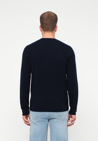 Bărbat întors cu spatele, purtând un pulover simplu, de culoare bleumarin închis, cu mâneci lungi, și blugi de culoare albastru deschis, pe un fundal alb.