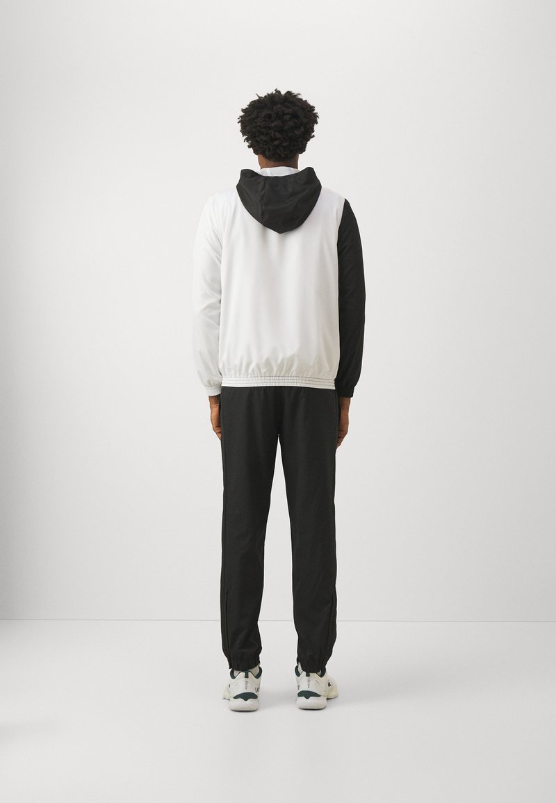 Giacca con cappuccio con maniche nere e corpo bianco, cintura elastica, abbinata a pantaloni neri e scarpe da ginnastica, vista da dietro.