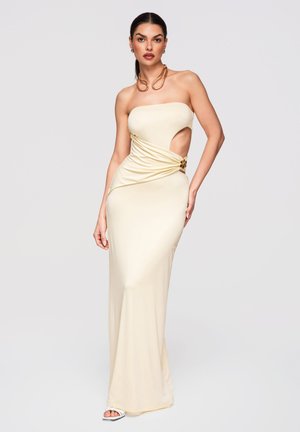 Kobieta o długich, ciemnych włosach, ubrana w kremową, bezramienną suknię wieczorową z wycięciem z tyłu, nosząca złote kolczyki koła i naszyjnik, stojąca na jednolitym tle.