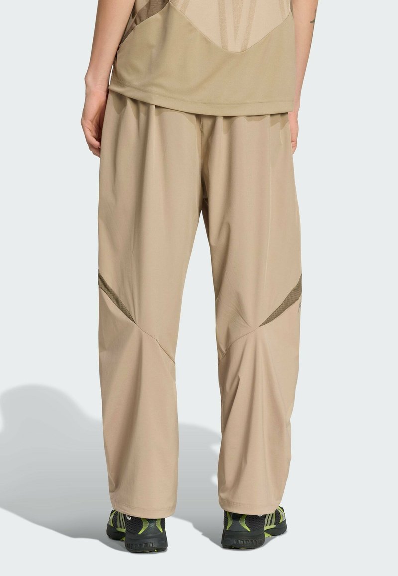 Un pantalon de sport beige présente une coupe ample, des panneaux texturés et des accents contrastants près des genoux, associé à des baskets noires et vertes.