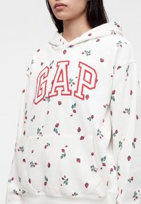 Personne portant un sweat à capuche blanc avec le logo rouge GAP et de petits motifs fraise, poche avant visible, tête partiellement visible sans yeux.