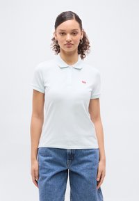 Νεαρή γυναίκα με κατ σαρά μαλλιά, φορώντας κρίκους στα αυτιά, ένα ανοιχτό μπλε πόλο Levi’s και μπλε τζιν, στέκεται μπροστά σε ένα απλό λευκό φόντο.