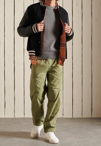 Mann in schwarzer Collegejacke, grauem Pullover, orange kariertem Hemd, olivgrünen Cargo-Hosen und weißen Sneakers, der vor einer weißen Holztafel steht.