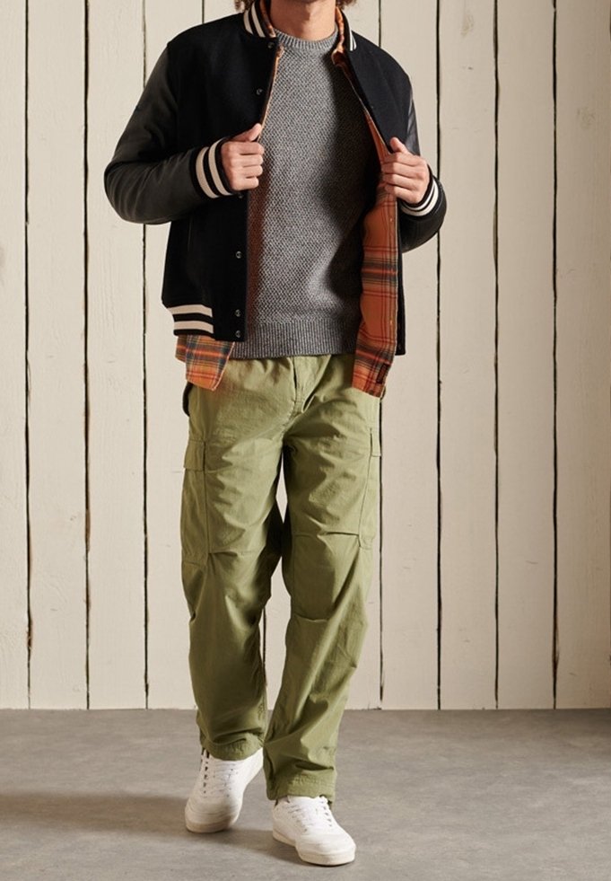 Mann in schwarzer Collegejacke, grauem Pullover, orange kariertem Hemd, olivgrünen Cargo-Hosen und weißen Sneakers, der vor einer weißen Holztafel steht.