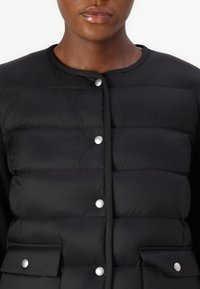 Zwarte gewatteerde vest met een quilted design, ronde halslijn en grote zilveren drukknoopsluitingen. Voorzien van twee voorkeurszakken met kleppen. Textuurstof.