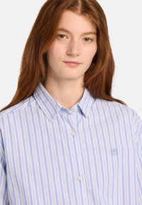 Jeune femme aux longs cheveux roux portant une chemise à rayures bleues et blanches avec un petit logo brodé sur la poitrine.