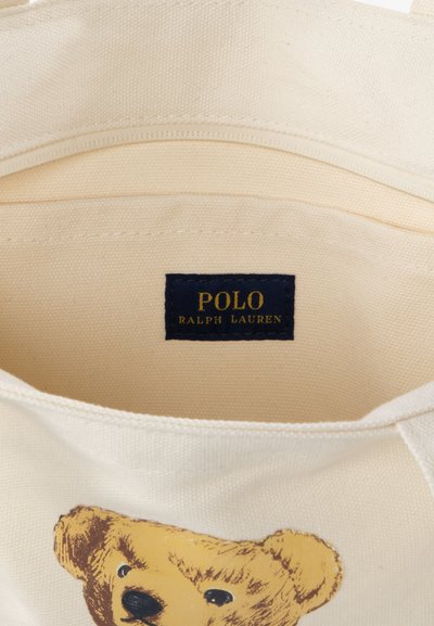 Geantă din material bej cu o etichetă neagră cusută pe care scrie „POLO RALPH LAUREN” și un chip de ursuleț imprimat aproape de partea de jos.