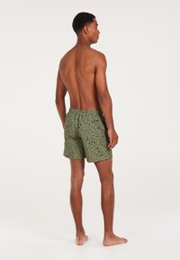 Zielone spodenki kąpielowe z czarnym, abstrakcyjnym wzorem, elastycznym paskiem w talii i jedną tylnią kieszenią. Lekka, teksturowana tkanina.