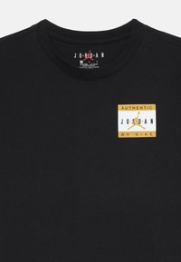T-shirt nera a girocollo con un piccolo logo dorato e bianco "Authentic Jordan by Nike" sul lato sinistro del petto.