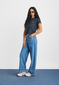 Svart avkortet t-skjorte med hvit logo, lyseblå vide denimjeans og lys rosa sneakers. Modellen har på seg mørke solbriller.