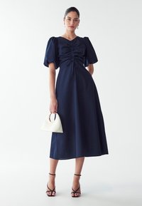 Marineblå midikjole med puffærmer og indsamlet detaljer, parret med en hvid håndtaske og strappy sorte sandaler.