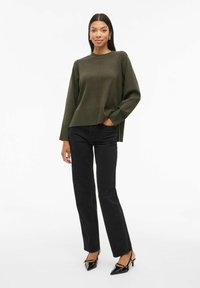 Maglione a maglia verde oliva con un design testurizzato, abbinato a jeans neri a gamba dritta e decolletè neri a punta. Silhouette semplice e moderna.