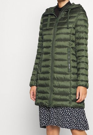Personne portant une veste à capuche matelassée verte avec fermeture éclair à l’avant et poches zippées sur une jupe fleurie noire et blanche.