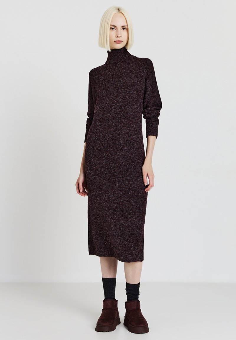 Abito in maglia di un marrone scuro con collo alto, maniche lunghe e silhouette aderente. Indossato con calze scure e stivaletti alla caviglia in suede marroni.