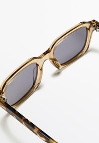 Sunglasses med en ljusbrun plastbåge, grå linser och mönstrade mörkbruna skalmar. Rektangulär form med en accentuerad ögonbrynsdel.