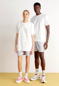 To modeller står side om side. Kvinden har en hvid T-shirt og stribede shorts på; manden bærer en matchende hvid T-shirt og stribede shorts. Begge har atletiske sko på.