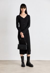 Abito lungo nero a maniche lunghe con bottoni, dotato di scollatura profonda e spacco laterale. Abbinato a stivali neri chunky sopra la caviglia e a una piccola borsa nera.